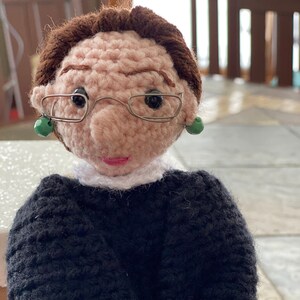Puede incluir: Mu&ntilde;eca de crochet con cabello casta&ntilde;o, que lleva gafas negras y una t&uacute;nica negra con cuello blanco. La mu&ntilde;eca tiene pendientes verdes y una cara rosa.