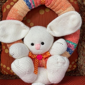 May include: A white crocheted bunny rabbit with orange and white polka dot ribbon sits in front of a crocheted wreath with orange, pink, and blue stripes.