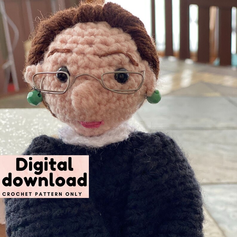 Puede incluir: Descarga de patr&oacute;n de crochet para una mu&ntilde;eca de crochet de Ruth Bader Ginsburg. La mu&ntilde;eca lleva una t&uacute;nica negra, un cuello blanco y gafas. La mu&ntilde;eca tiene el pelo casta&ntilde;o y pendientes verdes. El texto "Descarga digital" y "Patr&oacute;n de crochet solamente" est&aacute; sobre un fondo rosa.