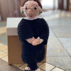 Puede incluir: Mu&ntilde;eca de crochet de una mujer con una t&uacute;nica negra, gafas y pendientes. La mu&ntilde;eca est&aacute; de pie con las manos juntas delante de ella.