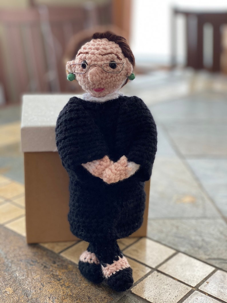 Puede incluir: Mu&ntilde;eca de crochet de una mujer con una t&uacute;nica negra, gafas y pendientes. La mu&ntilde;eca est&aacute; de pie con las manos juntas delante de ella.