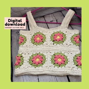 Könnte beinhalten: Ein Häkeltop mit weißem Hintergrund und grünen und rosa Blumenmotiven. Das Top hat eine quadratische Form und ist mit einem Granny-Square-Stich gearbeitet. Das Top hat dünne Träger und hängt an einem rosa Kleiderbügel.