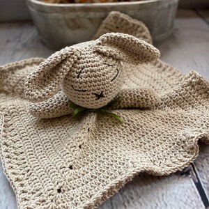 May include: A crocheted beige bunny security blanket with a scalloped edge. The bunny has a green ribbon around its neck.