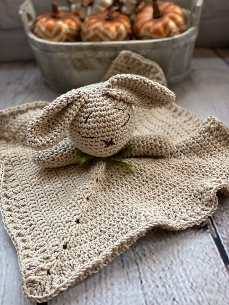 May include: A crocheted beige bunny security blanket with a scalloped edge. The bunny has a green ribbon around its neck.