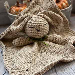 May include: A crocheted beige bunny security blanket with a green bow. The blanket is made with a soft, textured yarn and features a sweet bunny face with embroidered eyes and a stitched nose.