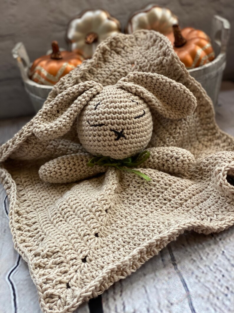 May include: A crocheted beige bunny security blanket with a green bow. The blanket is made with a soft, textured yarn and features a sweet bunny face with embroidered eyes and a stitched nose.