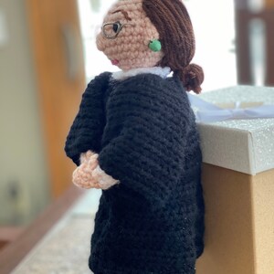 Puede incluir: Mu&ntilde;eca de crochet de una mujer con una t&uacute;nica negra y gafas con un pendiente verde. La mu&ntilde;eca est&aacute; de pie junto a una caja blanca y marr&oacute;n.