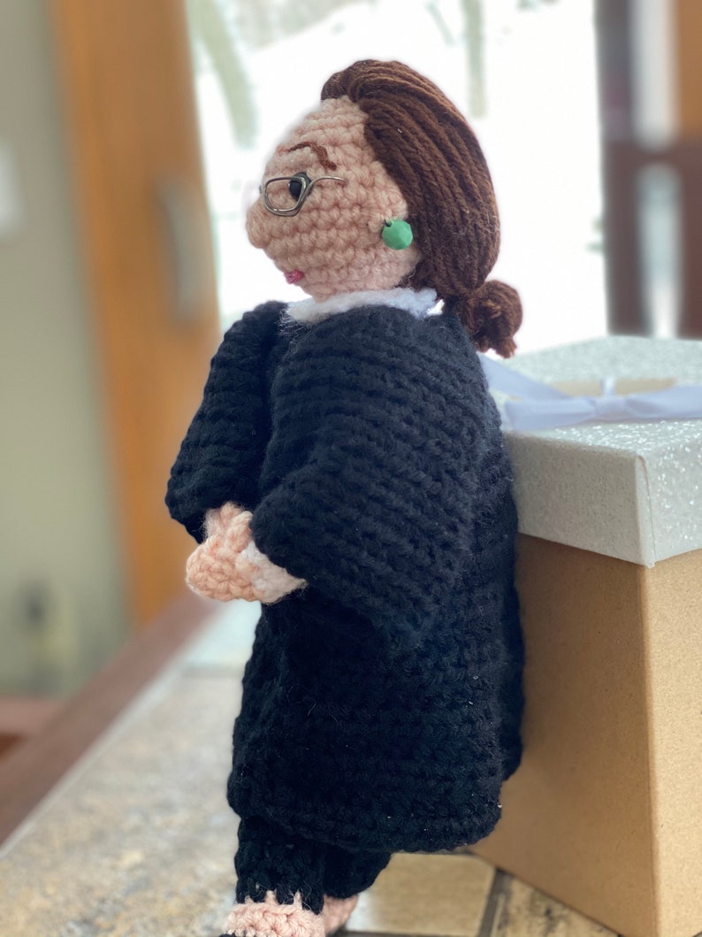 Puede incluir: Mu&ntilde;eca de crochet de una mujer con una t&uacute;nica negra y gafas con un pendiente verde. La mu&ntilde;eca est&aacute; de pie junto a una caja blanca y marr&oacute;n.