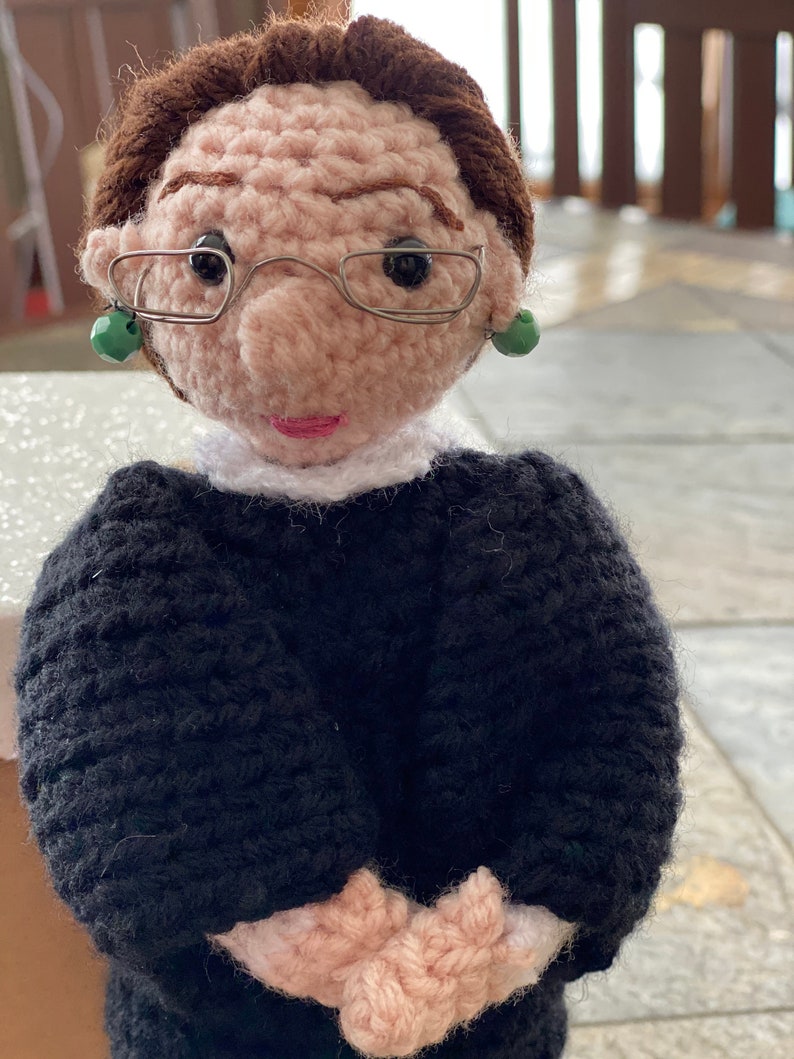 Puede incluir: Mu&ntilde;eca de crochet con cabello casta&ntilde;o, que lleva gafas negras y una t&uacute;nica negra con cuello blanco. La mu&ntilde;eca tiene pendientes verdes y una cara rosa.
