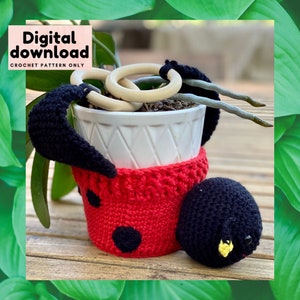Puede incluir: Una maceta de ladybug de ganchillo con detalles en negro y rojo. La maceta contiene una pequeña planta con anillos de madera. La maceta está sentada en una mesa de madera.