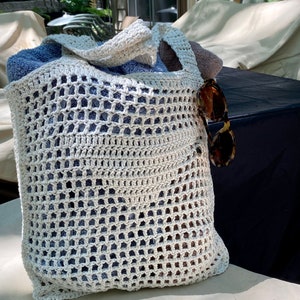 May include: A white crocheted tote bag with a blue towel inside. The bag is sitting on a white surface with a black surface behind it.