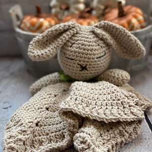 May include: A crocheted beige bunny lovey with a blanket attached. The bunny has a stitched-on face with black eyes and a black nose.