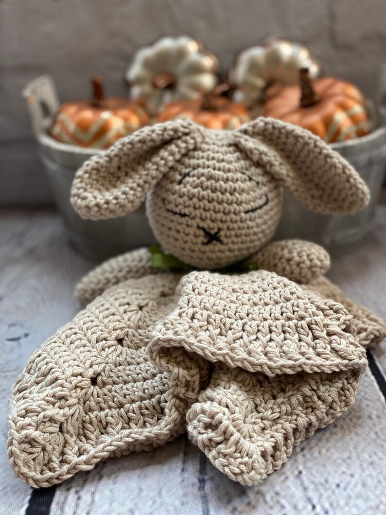 May include: A crocheted beige bunny lovey with a blanket attached. The bunny has a stitched-on face with black eyes and a black nose.