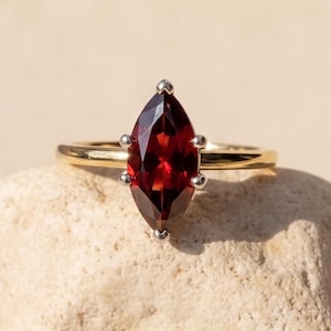 May include: A gold ring featuring a marquise-cut deep red garnet gemstone held in a six-prong silver setting. The band is a smooth, polished yellow gold, and the faceted stone is positioned vertically, showcasing its dark crimson color and sharp points.