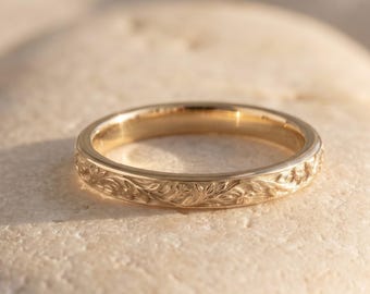 Può includere: Un anello in oro caratterizzato da un motivo floreale inciso e dettagliato che avvolge l'intera fascia esterna. Il metallo presenta una finitura lucida e riflettente che cattura la luce, mettendo in risalto gli intricati motivi a foglie e viticci.