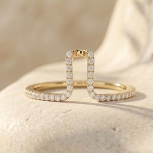 May include: A gold ring with a unique open design. The ring features a rectangular centerpiece and a band, both encrusted with small, sparkling white gemstones. The ring is set against a soft, neutral background, highlighting its elegant details.