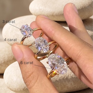 May include: Three gold rings with oval-cut diamond center stones, labeled as 3, 4, and 6 carats. The rings are displayed on a hand, showcasing the varying sizes of the diamonds. The rings have a classic, elegant design.