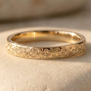 May include: A gold ring featuring a continuous engraved floral pattern around the entire outer band. The metal has a polished, reflective finish that catches the light, highlighting the intricate details of the small flowers and vines.