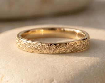 Può includere: Un anello in oro caratterizzato da un motivo floreale inciso in modo continuo lungo tutta la fascia esterna. Il metallo presenta una finitura lucida e riflettente che cattura la luce, mettendo in risalto i dettagli dei fiori e dei tralci.