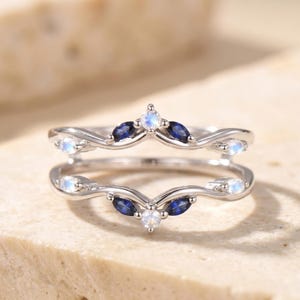 May include: Two silver rings with a wave design, each adorned with dark blue and white gemstones. The rings are stacked, creating a layered effect. The gemstones are in a leaf-like shape.