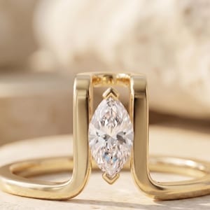 May include: A gold ring featuring a marquise-cut diamond. The diamond is set between two vertical gold bars, with a curved band that wraps around the finger. The ring is displayed on a neutral surface, highlighting its elegant design.