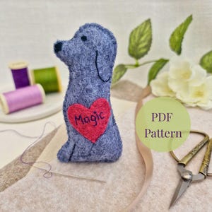 Può includere: Un cane in feltro fatto a mano in tonalità blu con un cuore rosso con la scritta "Magic". Il cane è seduto su una superficie beige, con un ago e filo nelle vicinanze. L'immagine include anche rocchetti di filo e un'etichetta "PDF Pattern".