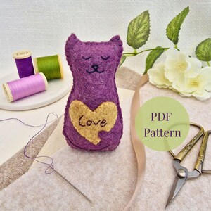 Può includere: Un gatto di feltro viola fatto a mano con un cuore dorato con la scritta "Love". Il gatto ha gli occhi chiusi ed è circondato da materiali da cucito, tra cui rocchetti di filo, un ago e forbici. L'immagine include anche una grafica "PDF Pattern".