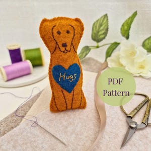 May include: A handmade felt dog in shades of orange with a blue heart that says "Hugs". The dog has embroidered details and is part of a PDF pattern. Sewing supplies, including thread and scissors, are in the background.