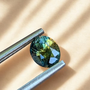 May include: An oval gemstone, displaying a blend of blue, green, and yellow tones, is held by silver tweezers. The facets of the gemstone catch the light, producing a sparkling effect. The background is a soft, neutral colour.
