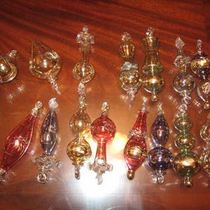 Könnte beinhalten: Eine Sammlung verzierter Glasornamente in verschiedenen Formen und Farben, darunter Gold, Rot und Blau. Die Ornamente sind auf einer reflektierenden braunen Oberfläche angeordnet, die ihre komplizierten Designs hervorhebt.