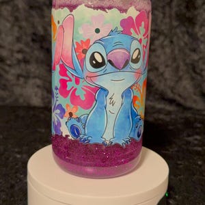 May include: A clear, cylindrical tumbler featuring a cartoon illustration of Stitch from Lilo & Stitch. The design includes Stitch in blue, surrounded by colorful flowers. The top and bottom sections are filled with purple glitter. The tumbler sits on a white rotating display stand.