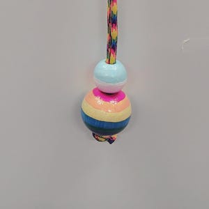May include: A hanging ornament featuring a multicolored braided cord with a small light blue bead above a larger spherical bead painted with horizontal stripes in pink, peach, yellow, blue, and green, ending in a knotted cord detail.