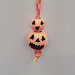 May include: A Halloween-themed ornament featuring two stacked, peach-colored, round beads painted to resemble jack-o'-lanterns. The faces are black with white accents. The ornament hangs from a pink, yellow, and brown braided cord.