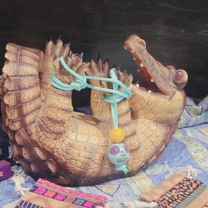 May include: A detailed, brown alligator-shaped decorative planter with an open mouth, featuring textured scales. A turquoise cord is tied around the alligator's body, with a decorative bead and skull charm hanging from it. The planter is set on a patterned fabric.