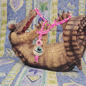 May include: A brown alligator figurine with an open mouth, holding a pink and white beaded necklace with two eyeball charms. The alligator is lying on its back. The background is a blue and green patterned fabric.