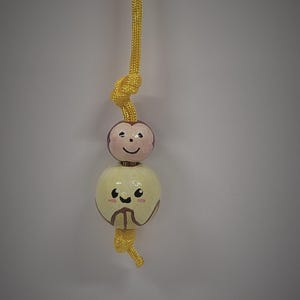 May include: A yellow cord hanging vertically with two round wooden beads attached. The top bead is painted with a pink monkey face, while the larger bottom bead is painted with a cream-colored face featuring dark eyes, a smile, and brown markings.
