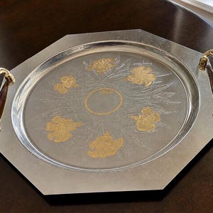 May include: A silver-toned octagonal serving tray featuring a central circular gold-toned ring surrounded by five gold-toned floral motifs. The tray includes two curved handles with a brown wood-grain pattern and gold-toned metal accents.