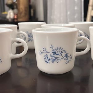 May include: A collection of six white ceramic mugs, each adorned with a blue floral motif. The mugs feature a rounded shape, a curved handle, and a blue floral design. They are displayed on a dark surface.