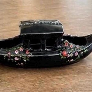 May include: A black ceramic gondola-shaped planter or decorative vessel featuring a central cabin structure and hand-painted floral details in shades of pink, red, and green on the exterior hull. The piece has a glossy finish and curved ends.