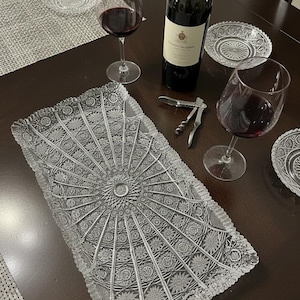 May include: A rectangular, clear glass serving tray with a detailed, starburst pattern. The tray is surrounded by wine glasses filled with red wine, a bottle of wine, and small glass plates. A wine opener is also present.