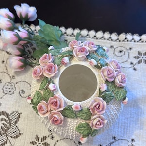 May include: A white ceramic vase with a circular opening, decorated with pink rose blossoms and green leaves. The vase has a textured, dotted surface. Artificial pink roses are placed next to the vase.