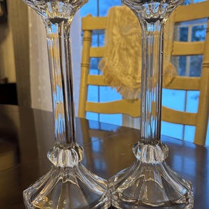 May include: A pair of clear glass candlesticks with a fluted stem and a hexagonal base. The candlesticks are approximately 25 cm tall. The candle holders have a flared top to hold a candle. The background includes a wooden table and a window.