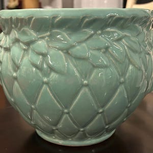 May include: A round, light teal ceramic planter with a textured, quilted pattern. The rim features a leaf-like design. The planter is suitable for indoor or outdoor use and adds a touch of elegance to any space.
