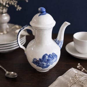 May include: A white porcelain teapot featuring a rounded body, a curved spout, and a lid with a small knob. The teapot is decorated with hand-painted blue floral motifs on the body, handle, spout, and lid, showcasing a glossy, smooth finish.