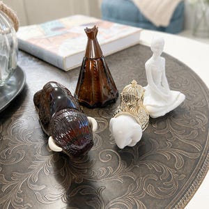 May include: A collection of four decorative figurines including a dark brown glass bison, a conical brown glass bottle, a white ceramic elephant with a gold-toned ornate saddle, and a white ceramic seated figure in a meditative pose.