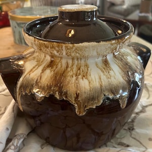 May include: A brown ceramic bean pot with a lid. The pot has a glossy finish with a two-tone design, featuring a dark brown base and a lighter, dripped glaze effect. The lid is also brown with a gold rim.