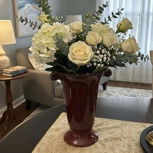 May include: A burgundy vase filled with a bouquet of white roses, hydrangeas, and greenery. The vase has a flared top and a pedestal base. The arrangement is set on a table with a gold-colored runner, creating a classic floral display.