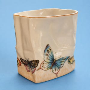 May include: A cream-colored ceramic vase with a crinkled, paper-bag-like texture. The exterior features hand-painted butterflies in shades of blue, pink, and yellow, with a soft orange wash along the base and a thin orange rim at the top opening.