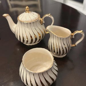 May include: A white and gold tea set comprising a teapot, creamer jug, and sugar bowl. The set has a white base with gold vertical stripes and gold detailing on the handles and rims. The teapot has a lid with a gold finial.