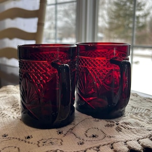May include: Two ruby red glass mugs with handles. The mugs feature a diamond and leaf pattern. They are placed on a cream lace tablecloth near a window.